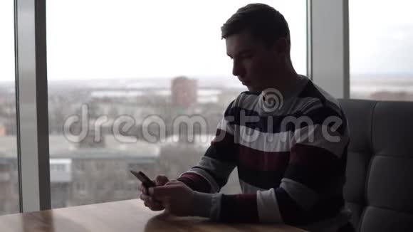一个带电话的年轻人坐在桌子旁一个男人坐在全景窗旁的咖啡馆里手里拿着一部智能手机视频的预览图