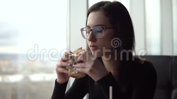 一个年轻的女人正在吃汉堡一个女孩坐在全景窗旁的咖啡馆里吃快餐视频的预览图