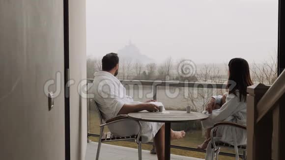 快乐的白种人夫妇穿着白色长袍喝着咖啡在房间露台上观看圣米歇尔山的景色视频的预览图
