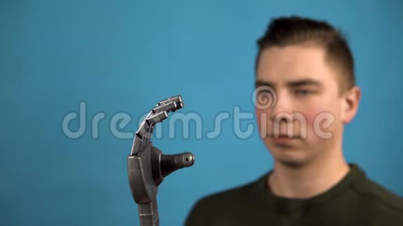 一个年轻人看着他的机械手臂赛博格在蓝色背景上用灰色的手挤压手指视频的预览图