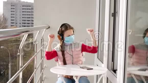 十几岁的女孩戴着面具和耳机在智能手机上听音乐在开放的阳台上春天阳光明媚的一天检疫视频的预览图