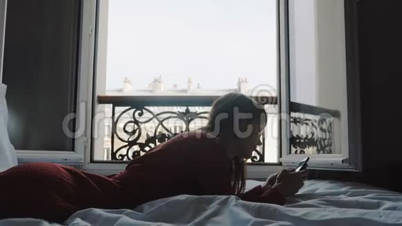 快乐成功的欧洲自由职业者女性在智能手机应用程序上在舒适的法国阳台床上打字慢动作视频的预览图