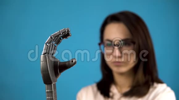 一个年轻的女人看着她的机械手臂赛博格在蓝色背景上用灰色的手挤压手指视频的预览图