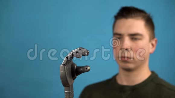 一个年轻人看着他的机械手臂赛博格在蓝色背景上用灰色的手挤压手指视频的预览图