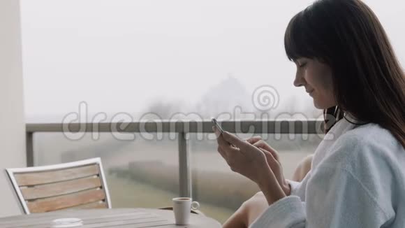 美丽的年轻微笑的女人使用智能手机应用程序在外面的酒店露台与惊人的雾秋景慢动作视频的预览图