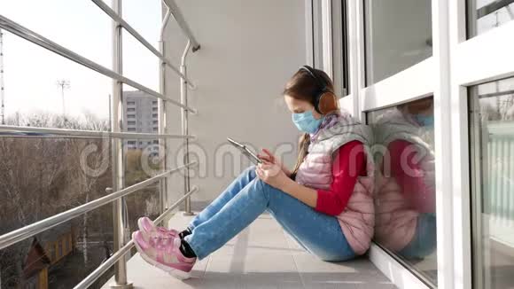十几岁的女孩戴着面具和耳机打字在平板电脑上工作在开放的阳台上春天阳光明媚的一天远程学习视频的预览图