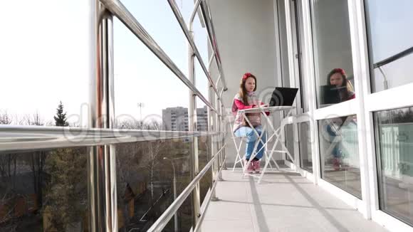 漂亮的少女正在打字在笔记本电脑上工作在开放的阳台上春天阳光明媚的一天远程学习概念视频的预览图