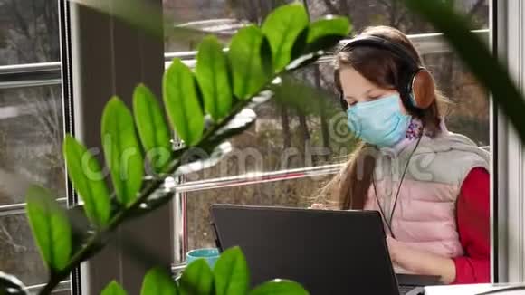 十几岁的女孩戴着面具和耳机在笔记本电脑上听音乐在网上聊天在开放的阳台上春天阳光明媚视频的预览图