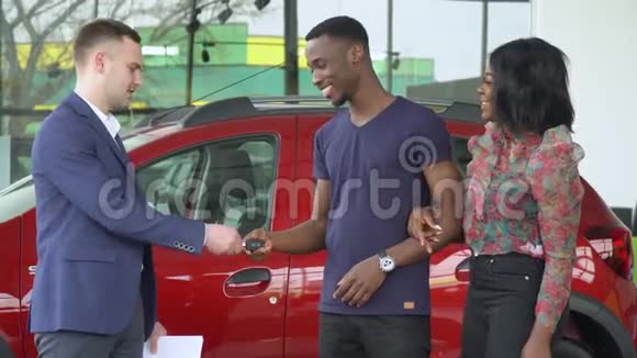 年轻的白种人英俊的汽车销售商给他的非裔美国顾客一把新车的钥匙视频的预览图