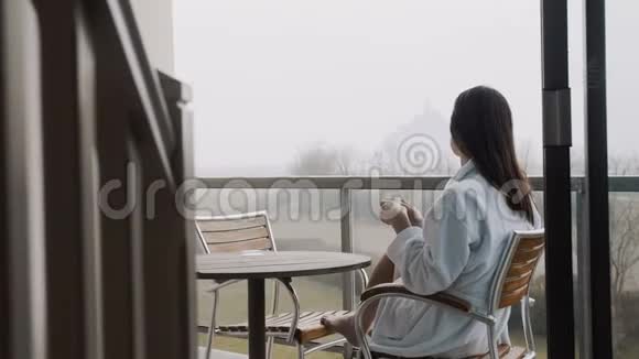 后景美丽的女士穿着晨衣在客房露台上享受咖啡带雾蒙圣米歇尔景观慢镜头视频的预览图
