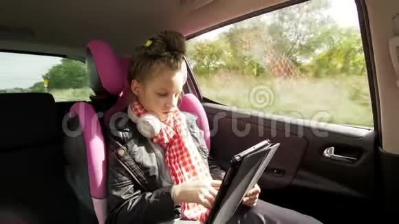 漂亮的小女孩带着耳机用平板电脑少年儿童坐在后座上带着皮带旅行视频的预览图