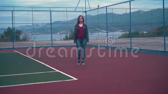 穿着自行车夹克和牛仔裤的漂亮而热情的女孩沿着篮球或网球场散步遥远的计划视频的预览图