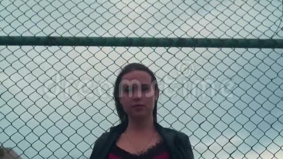 穿着自行车夹克的漂亮而热情的女孩站在铁栅栏的背景上视频的预览图
