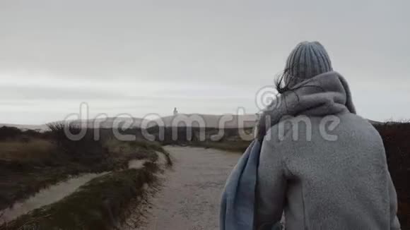 镜头跟随美丽的欧洲旅游女子穿着冬衣步行到远处阴云密布的海滩灯塔慢动作视频的预览图