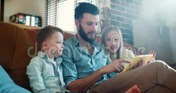 快乐的年轻白种人父亲用平板电脑读书给可爱的小儿子和女儿在家里沙发上慢动作视频的预览图