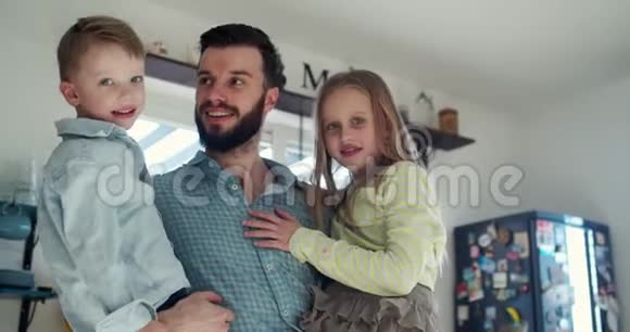 快乐的年轻白种人父亲的肖像看着镜头抱着小儿子和女儿在家里厨房慢动作视频的预览图
