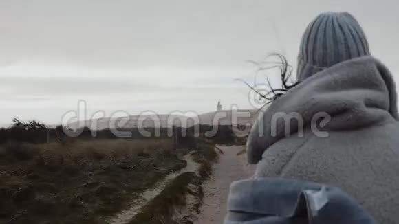 镜头跟随着一位穿着冬装的有思想的女游客走向遥远的阴暗海滩灯塔慢动作视频的预览图