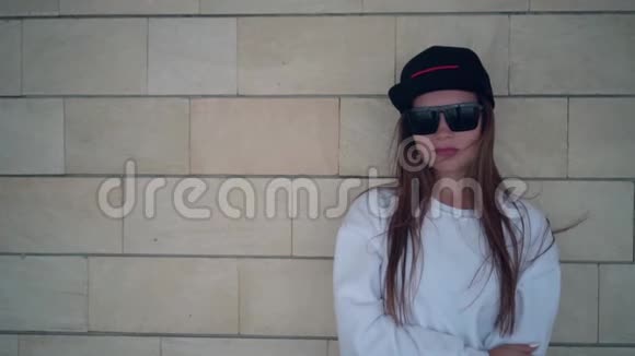 一个年轻性感的女孩戴着太阳镜长发穿着一件白色的运动衫站在米色的砖块墙上视频的预览图