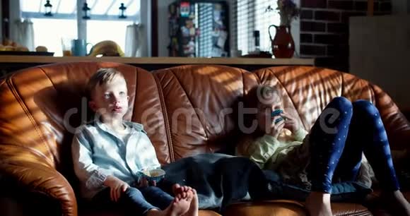 快乐的小可爱白种人哥哥姐姐孩子们在家看电视在沙发上吃松饼慢动作视频的预览图