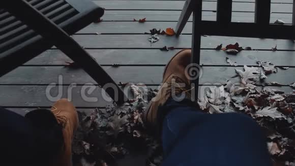 一个放松的人坐在秋天的露台上穿着温暖的棕色皮靴枯叶缓慢地飞翔视频的预览图