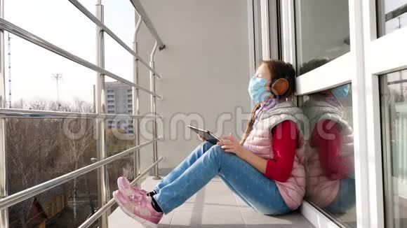 十几岁的女孩戴着面具和耳机在平板电脑上听音乐在开放的阳台上春天阳光明媚的一天隔离住宿视频的预览图