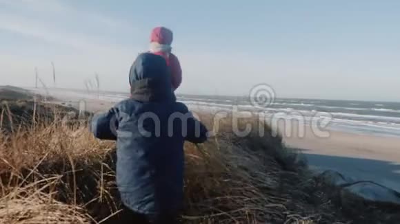 小女孩和男孩穿着温暖的外套在阳光明媚的冬天海滩缓慢的运动中沿着高大的草地沙丘行走视频的预览图