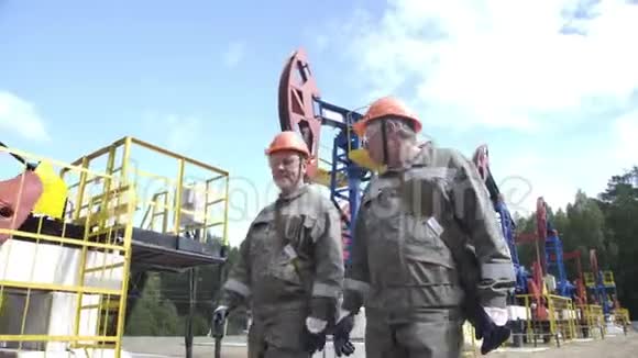 两名男性石油工人在油泵千斤顶附近行走和交谈石油工程师监督原油钻井平台视频的预览图