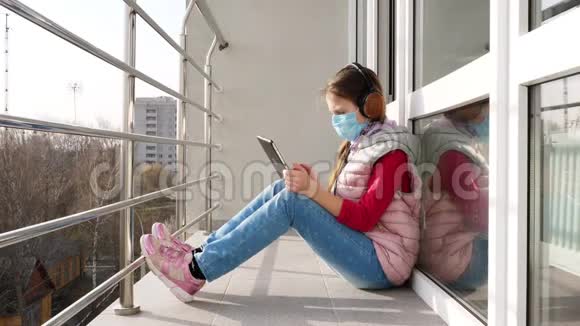 十几岁的女孩戴着面具和耳机打字在平板电脑上工作在开放的阳台上春天阳光明媚的一天远程学习视频的预览图