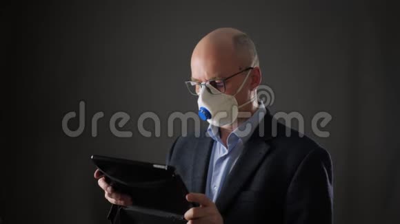 商人在防护面具使用平板电脑而病毒流行在黑色背景严肃的男人看着平板电脑视频的预览图