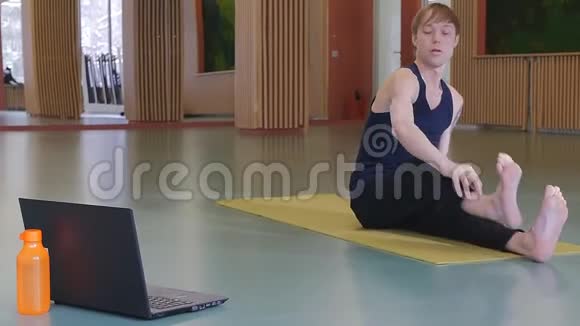 一个年轻可爱的家伙在网上教瑜伽坐在黄色垫子上做伸展运动工作室日光灯视频的预览图