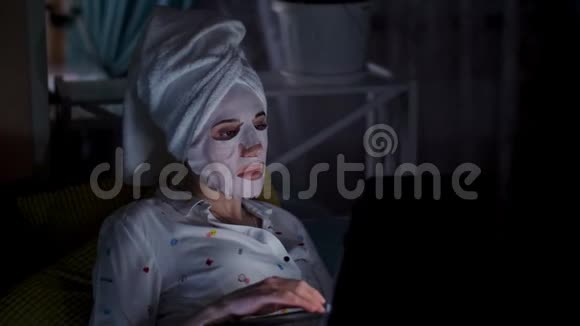 年轻女子戴着化妆面料的面具头上戴着白毛巾在笔记本电脑上工作晚上在屏幕上观看视频的预览图