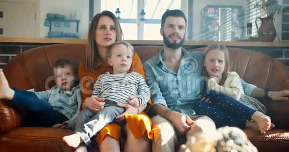 快乐的年轻白种人母亲父亲和三个可爱的孩子一起看电视周末在家里沙发慢动作视频的预览图