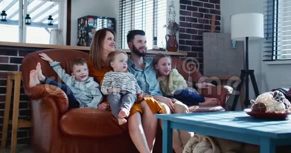 年轻快乐的欧洲母亲父亲和三个孩子微笑着在家一起在沙发上看有趣的电影慢动作视频的预览图