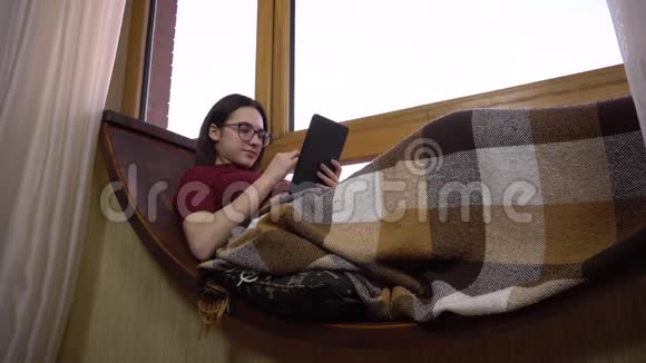 一位年轻女子正在平板电脑上发短信一个女孩躺在窗边的窗台上手里拿着一块平板电脑外部视频的预览图