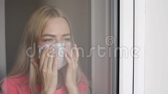 家庭隔离白人女人戴着医疗面具站在窗口向外看视频的预览图