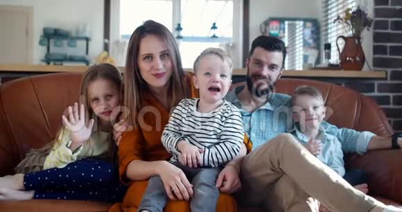 幸福的年轻家庭肖像白人父母和三个可爱的孩子看着相机在家里的沙发上慢动作视频的预览图