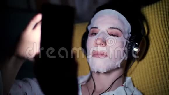 女人戴着白色化妆品面料面具带着耳机手里拿着智能手机手机听着看着看着视频的预览图