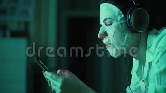 肖像年轻女子戴着白色化妆面料面具头上戴着耳机手里拿着智能手机手机视频的预览图