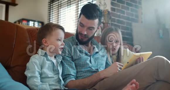 可爱的小白种人儿子和女儿在家里沙发上用平板电脑和父亲一起阅读和玩耍视频的预览图