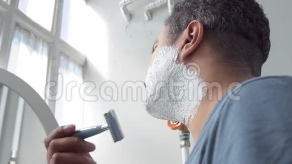 用泡沫侧视特写男人剃须然后相机移动在镜子中显示他的脸视频的预览图