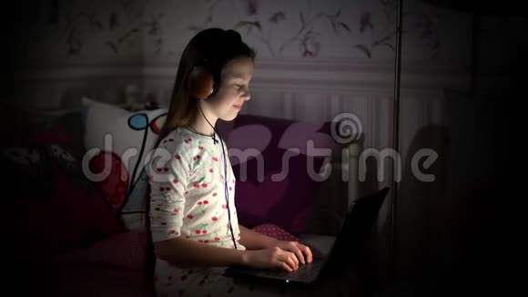 十几岁的女孩穿着睡衣戴着耳机看着笔记本电脑上的东西打字听音乐晚上在电脑里视频的预览图