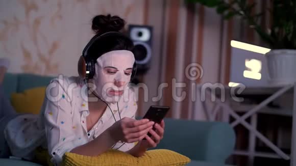 肖像年轻女子戴着白色化妆面料面具头上戴着耳机手里拿着智能手机手机视频的预览图