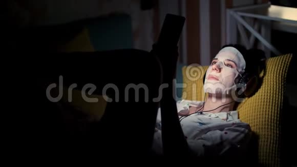 女人戴着白色化妆品面料面具带着耳机手里拿着智能手机手机听着看着看着视频的预览图