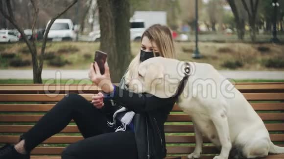 带着黑色面具的年轻女人坐在长凳上在公园的长凳上自拍在城市里视频的预览图