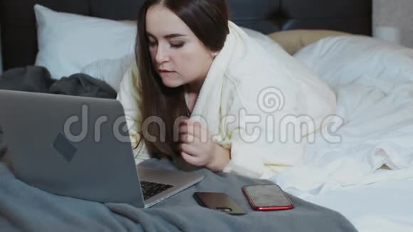 穿着浴袍的女人躺在床上用笔记本电脑和智能手机工作自由职业者在家工作在外面工作视频的预览图
