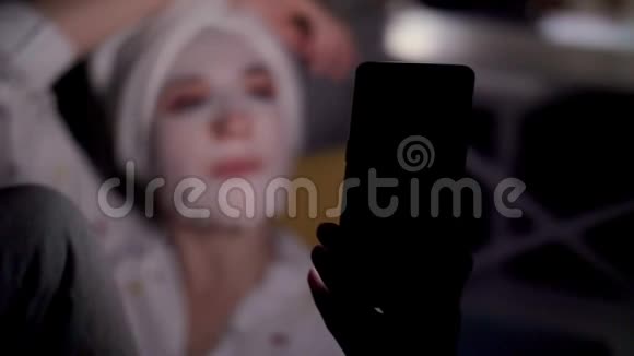 年轻女子戴着化妆面料面具头上戴着白色毛巾手里拿着智能手机听着看着晚上躺着视频的预览图