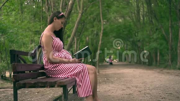一个怀孕的女孩坐在公园的长凳上一个穿着条纹白红色连衣裙的女孩热情地看书视频的预览图