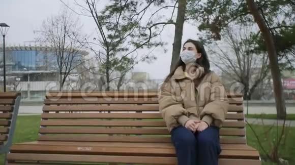 戴着口罩的漂亮女人坐在公园里的长凳上在冠状病毒COVID19流行时期视频的预览图