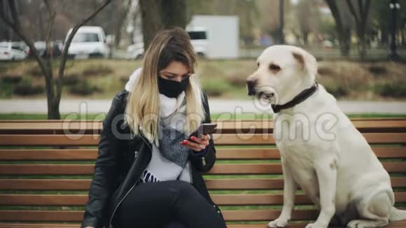带着黑色面具的年轻女子坐在长凳上在城市公园里使用电话视频的预览图