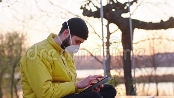 在公园里日落时分一个男人坐在椅子上时用电子平板电脑做特写他戴着一个防护面具视频的预览图